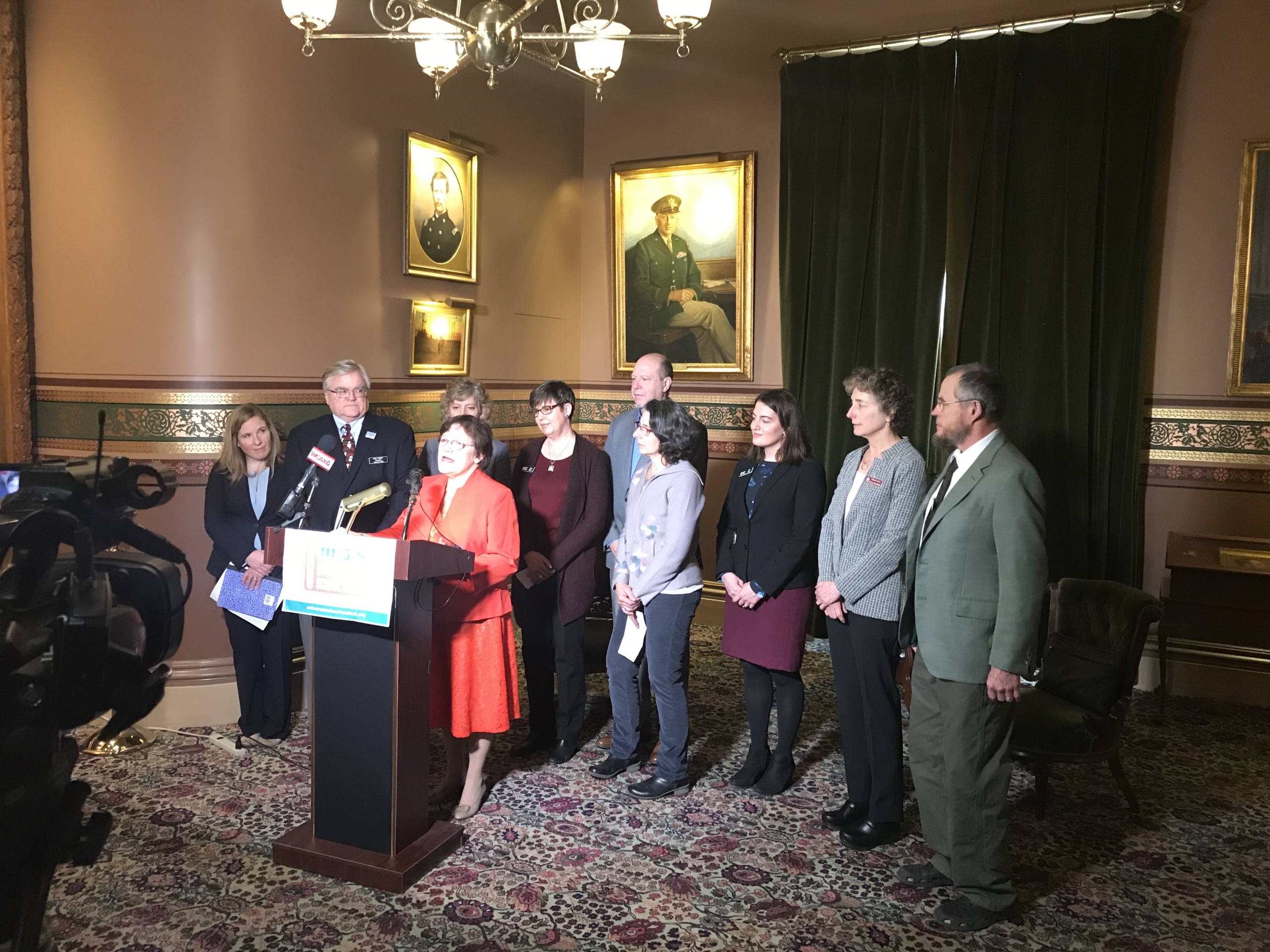 group of people standing behind a women at a podium giving a speech