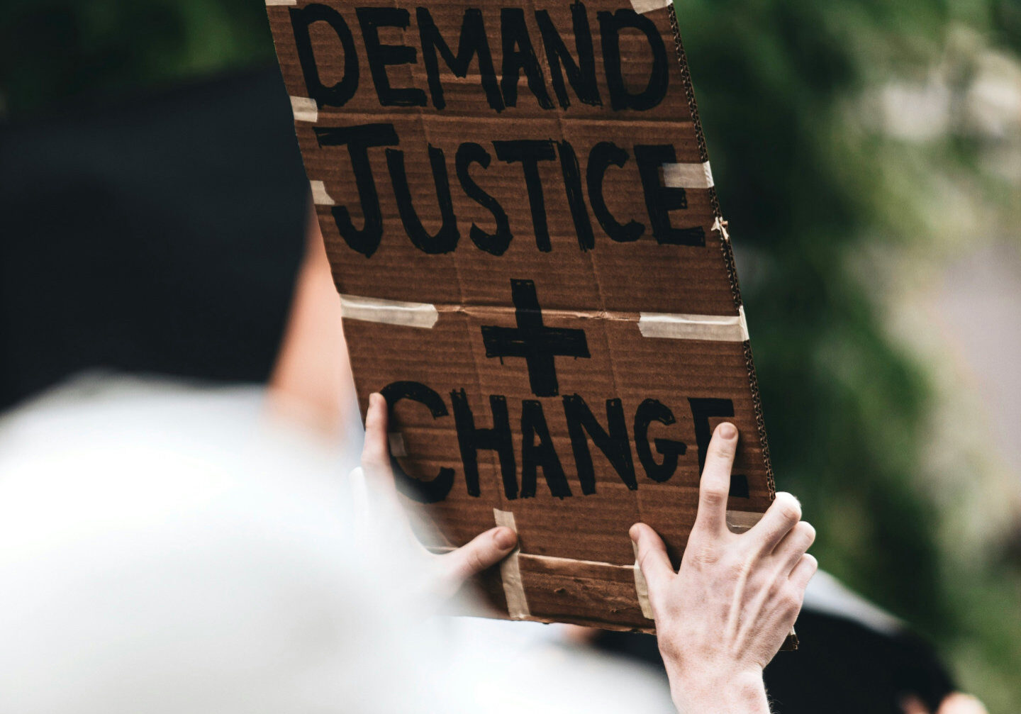 Person holding up a cardboard sign that reads: WE DEMAND JUSTICE + CHANGE.